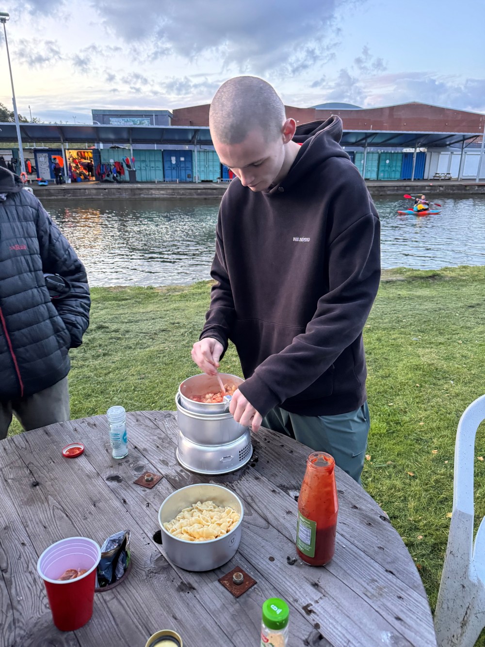 Person cooking pasta outdoors near a lake with kayaks in the background.