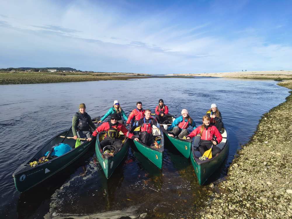 Group of eight people in canoes on a river, wearing life jackets, with a clear sky.