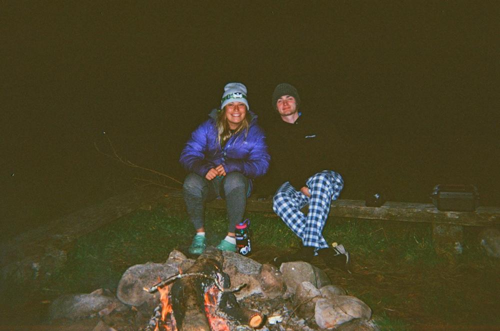 Two people sitting by a campfire at night, wearing warm clothes and hats.