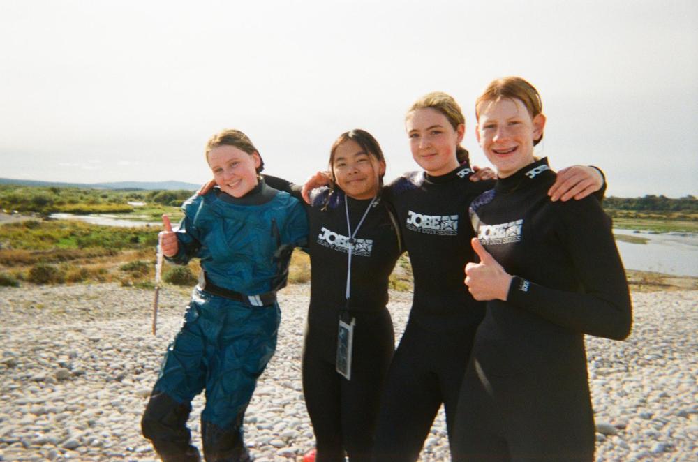 Four people in wetsuits smiling and giving thumbs up by a rocky riverbank.