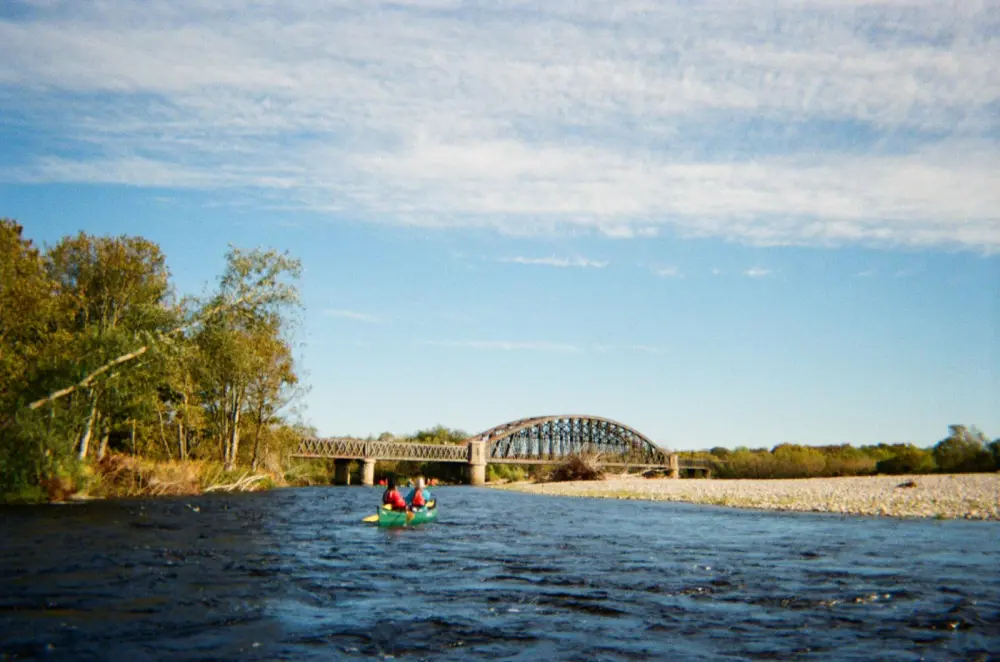 Two people kayaking on a river under a metal arch bridge on a sunny day.