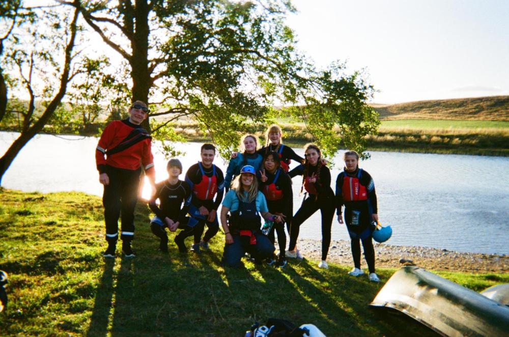 Group of people in life vests by a river, with trees and grassy landscape in the background.