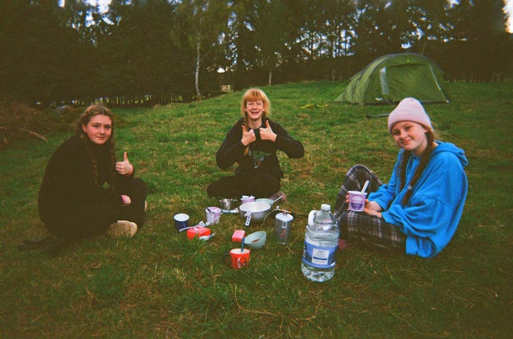 Three people sitting on grass near a tent, holding thumbs up and cups, with camping gear around.