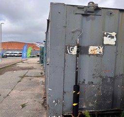 Rusty metal container with a hook on the side, on a deserted paved area under cloudy sky.