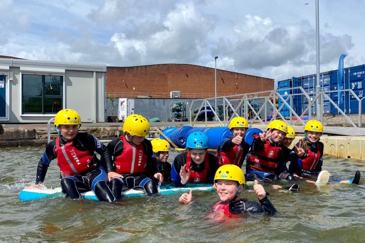Group of kids in helmets and wetsuits on paddleboards in a water sports area.