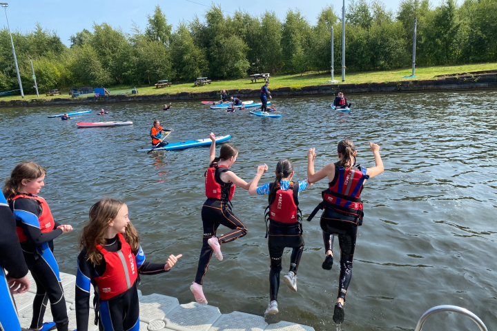 Children in life vests jump into a lake from a platform, with paddleboarders nearby.