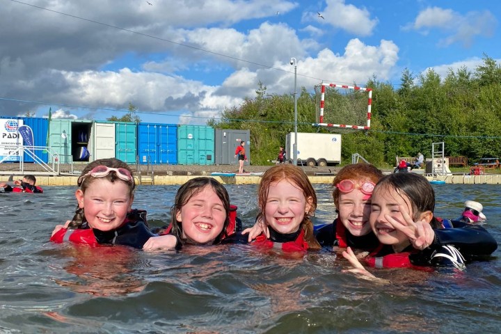 Five smiling children in water, wearing life vests, with a cloudy sky and storage containers in the background.