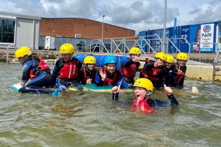Group of kids in helmets and wetsuits on a makeshift raft in a water sports setting.