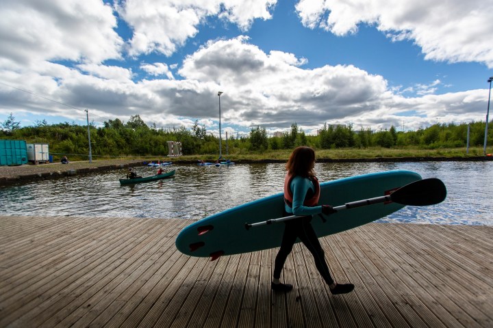 Person carrying paddleboard by a river under cloudy sky.