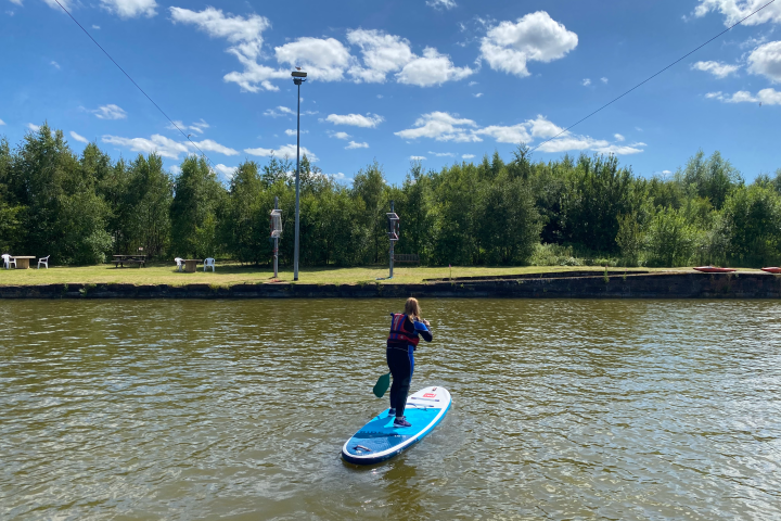Person paddleboarding on a river under a blue sky with trees in the background.