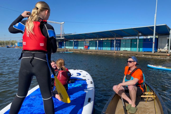 People on paddleboards and a boat on calm water, near a building on a sunny day.