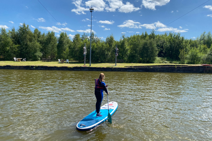 Person paddleboarding on a calm river under a blue sky with clouds.