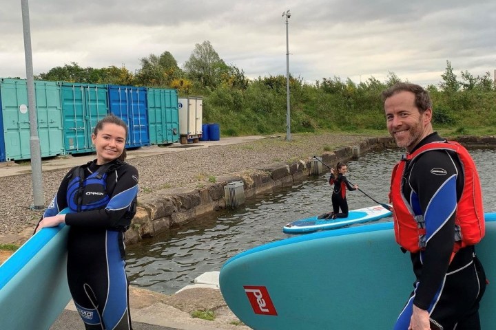 Two people in wetsuits holding paddleboards by the water.