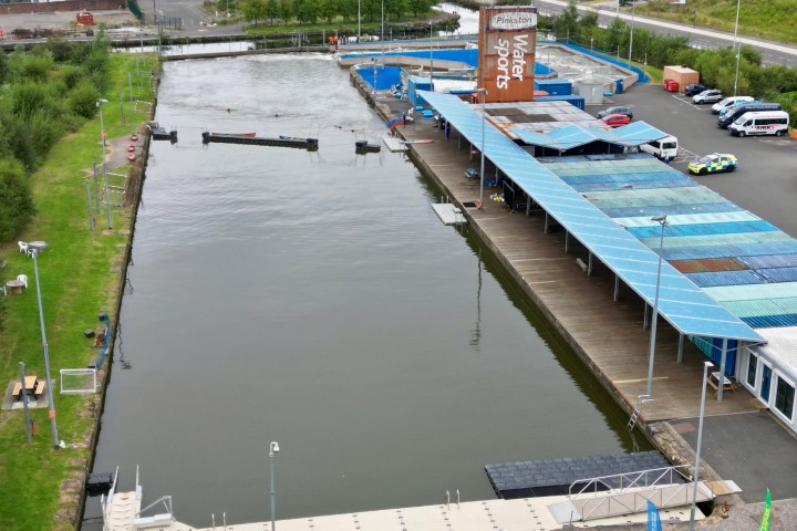 Aerial view of a watersports area with a long pool, blue-roofed building, and parked vans on the side.