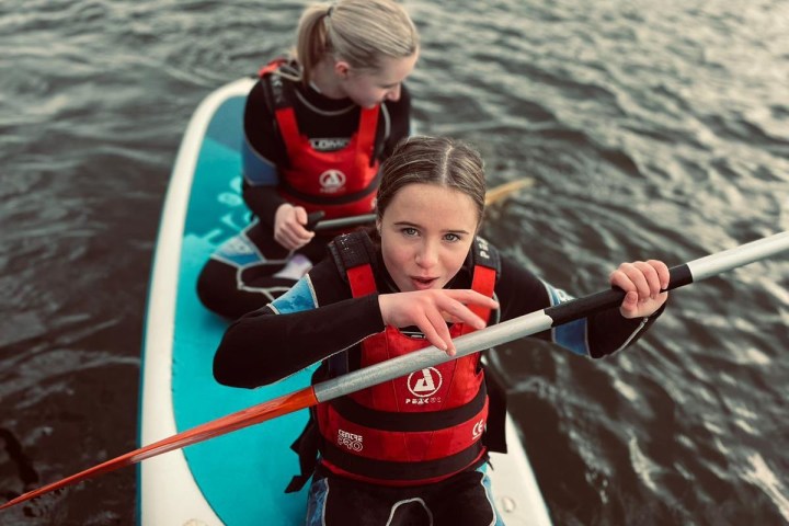 Two people in wetsuits on a paddleboard, one paddling on a lake.