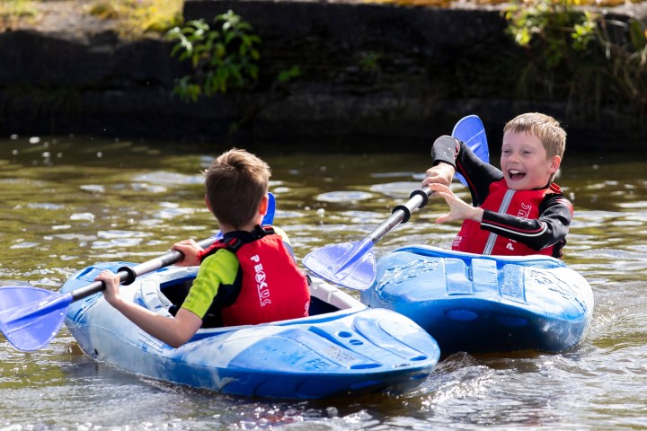 Two boys in red life vests kayaking on a sunny day.
