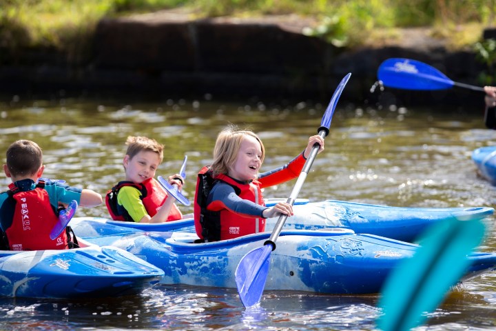 Children kayaking in blue kayaks on a sunny day, wearing red life jackets.