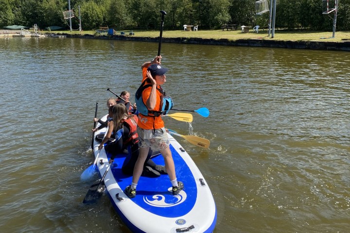 Four people kayaking on a large blue paddle board in a calm river.
