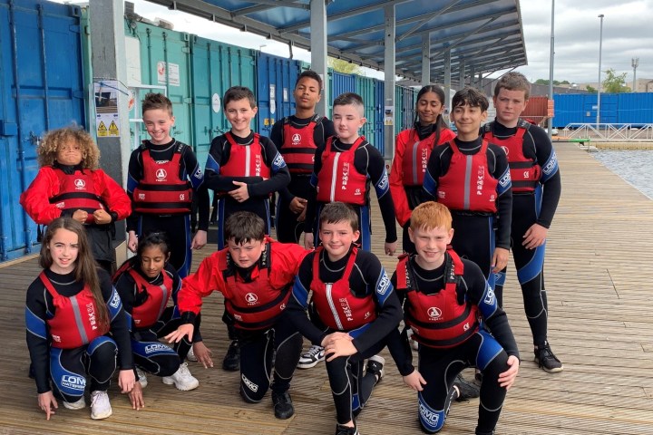 Group of kids in wetsuits and red life jackets posing on a wooden dock.