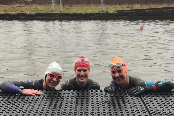 Three swimmers in wetsuits and swim caps smiling, holding onto a dock by a body of water.