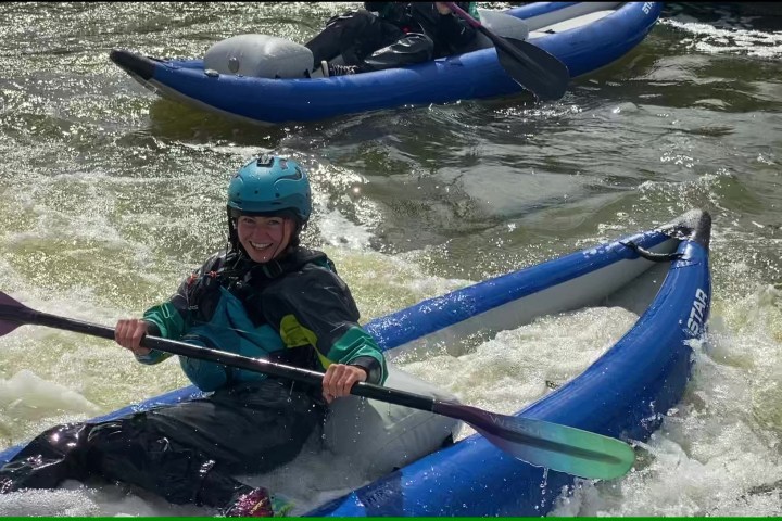 Person kayaking in rough water, smiling, wearing helmet and gear.