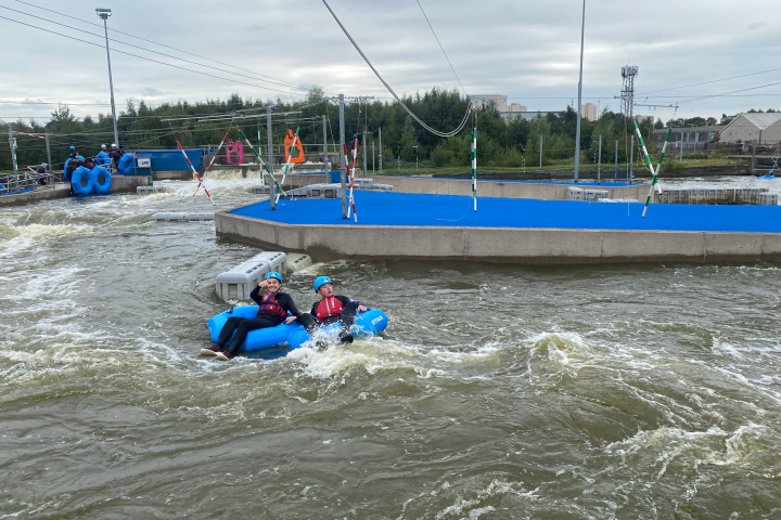 Two people in helmets white-water rafting on a course with blue barriers.