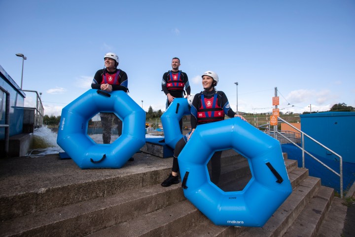 Three people in wetsuits with helmets holding blue inflatable rings on concrete steps.