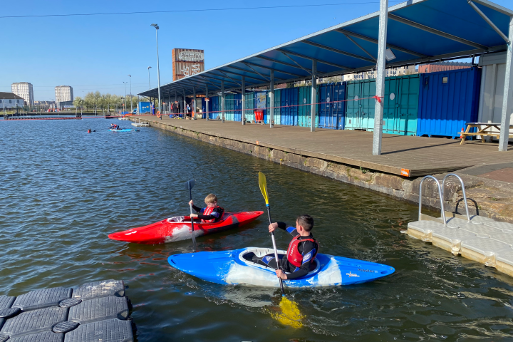 Two people kayaking near a dock at a waterside facility under a blue sky.
