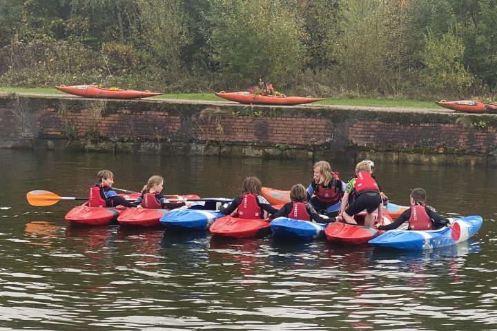Children in kayaks on a river near a grassy bank with trees.