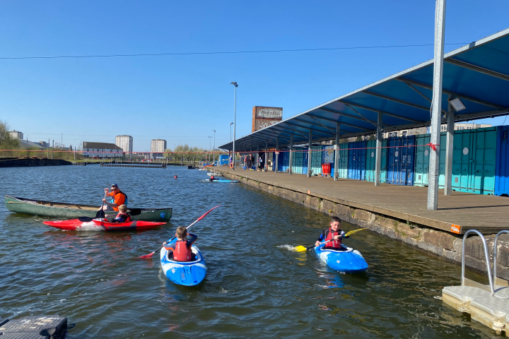People kayaking in a canal near a blue industrial building with a clear sky.