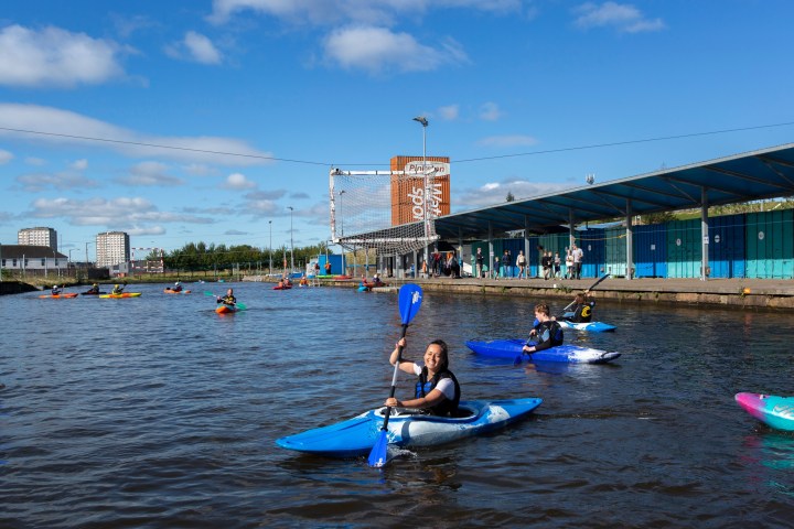 People kayaking on a river beside a blue covered walkway and urban buildings under a clear sky.