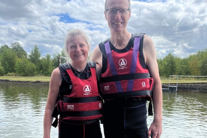 Two people in wetsuits and life vests by a lake, standing on a wooden deck.