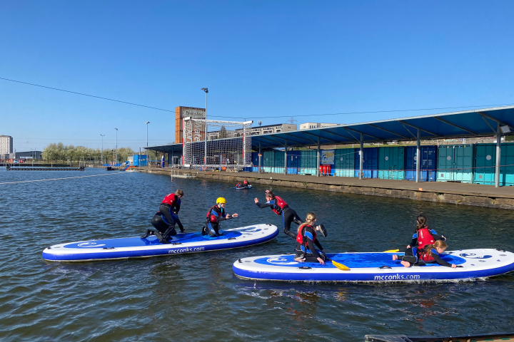 People in wetsuits on paddleboards in an urban waterway with buildings in the background.