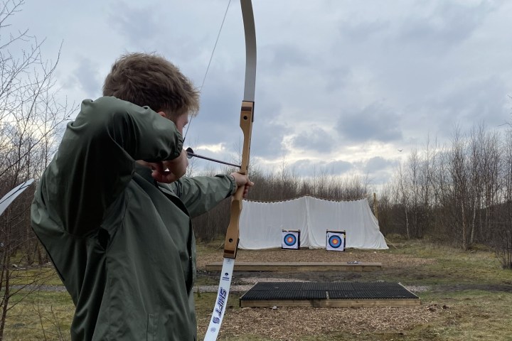 Person in green jacket aiming a bow at targets outdoors on a cloudy day.