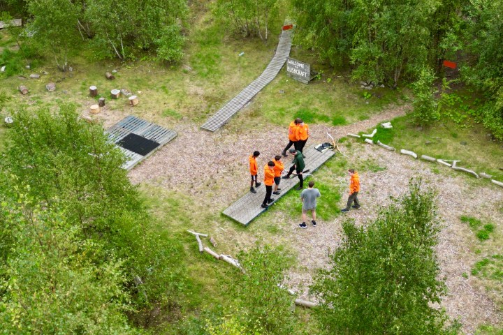 People in orange shirts balancing on wooden beams in a forest clearing.