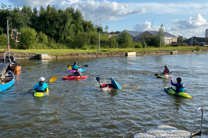 People kayaking on a river with trees and buildings in the background.
