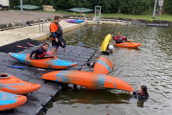 People in kayaks on a dock and in the water by a lake.