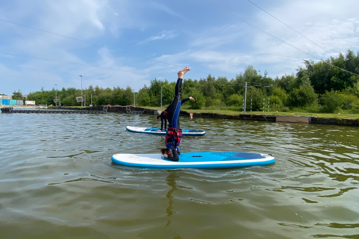 Two people doing headstands on paddleboards in a lake.