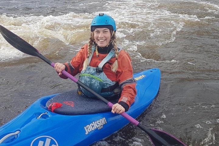 Smiling person in a kayak on a river wearing a helmet and life jacket.