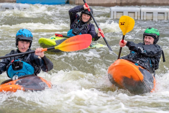 Three people kayaking on rough water, wearing helmets and life vests.