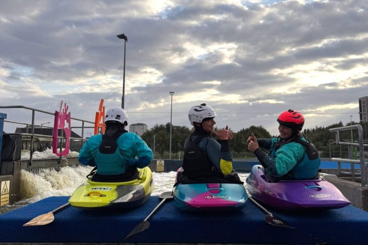 Three kayakers in helmets sit on a platform under a cloudy sky.