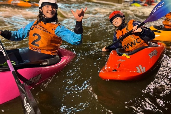 Two kayakers in helmets and vests smile and gesture, kayaking in an artificial whitewater course at night.