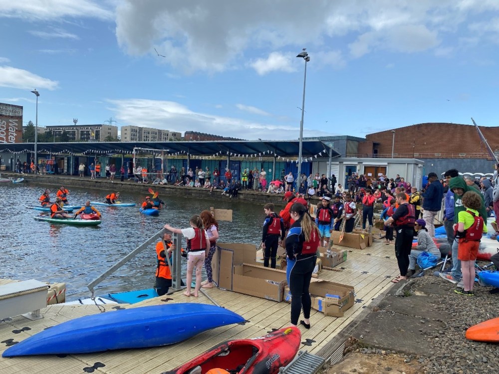Crowded pier with kayakers and onlookers near the water on a cloudy day.