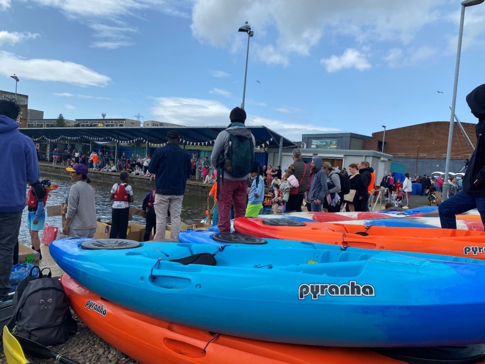 People gathering near stacked kayaks by a waterfront.