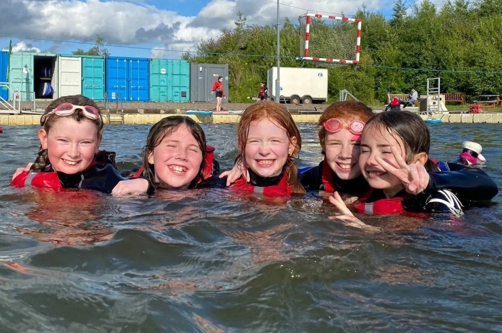 Five smiling children in water, wearing life vests and goggles, posing with peace signs, outdoor setting.