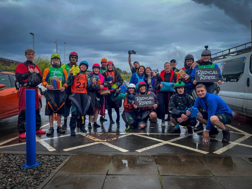 Group of kayakers in gear posing with smiles and 'Radical Ridez' signs on a wet pavement.